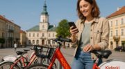 Dziewczyna wypożyczająca czerwono-biały rower z systemu Kielecki Rower Miejski za pomocą telefonu przy stacji w centrum Kielc. W tle Rynek, słoneczny dzień, miejski klimat.